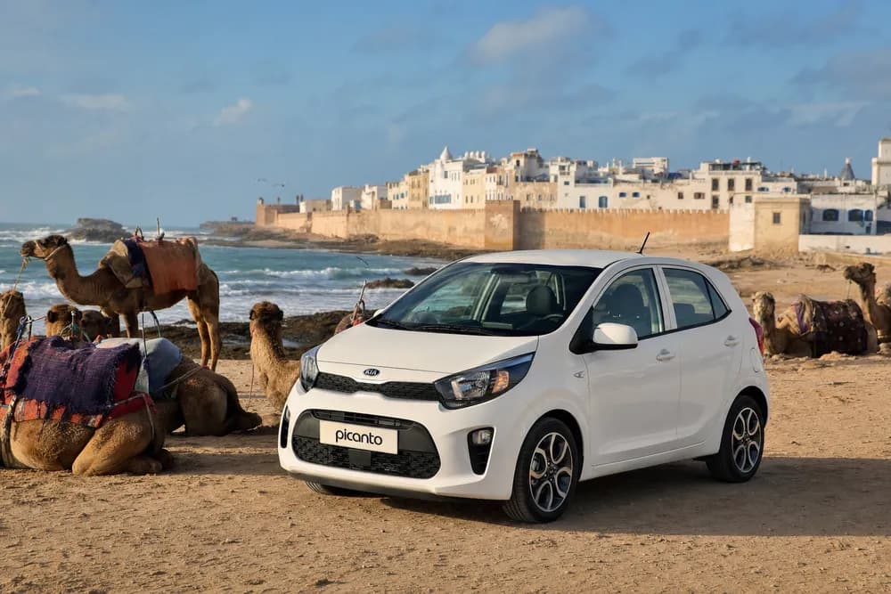 Coche de alquiler Kia Picanto en la playa de Essaouira con camellos y las murallas de la Medina de fondo